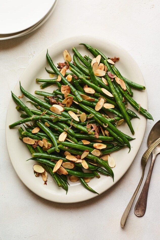 An overhead photo of a serving dish holding green beans almondine. A serving fork and spoon lay next to the dish.