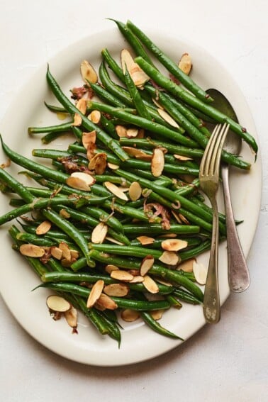 An overhead photo of a serving dish holding green beans almondine. A serving fork and spoon lay across the green beans..