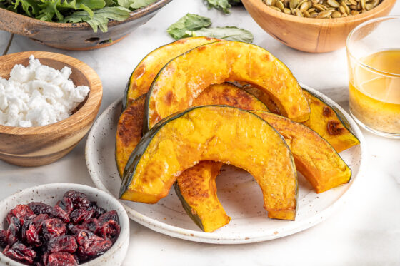 Freshly roasted kabocha squash slices piled on a plate. The plate is surrounded by other small bowls with cranberries and feta.