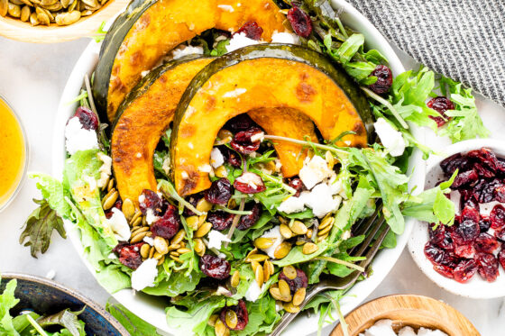 An overhead photo of a bowl of kale topped with 3 slices of roasted kabocha squash and scattered with dried cranberries, pumpkin seeds and feta. A fork rests in the bowl.