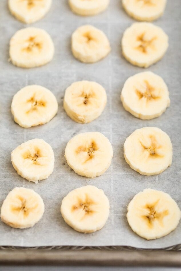 A metal sheet pan lined with parchment paper. There are banana slices across the parchment paper.