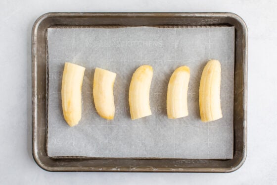 An overhead photo of a small sheet pan lined with parchment paper. There is a row of banana halves organized on the parchment paper.