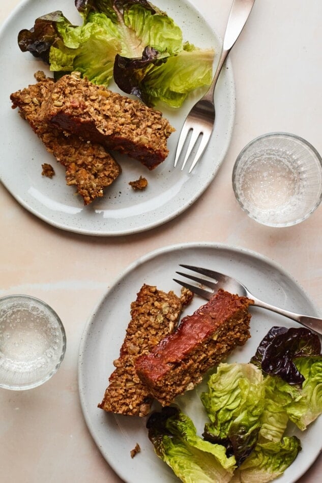 Two plates with two slices of lentil loaf and salad greens. Additionally, there are two forks and two glasses of water.