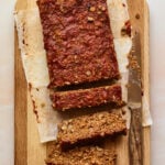 An overhead shot of a cutting board with a partially sliced lentil loaf on parchment paper. A knife lays on the cutting board perpendicular to the loaf.