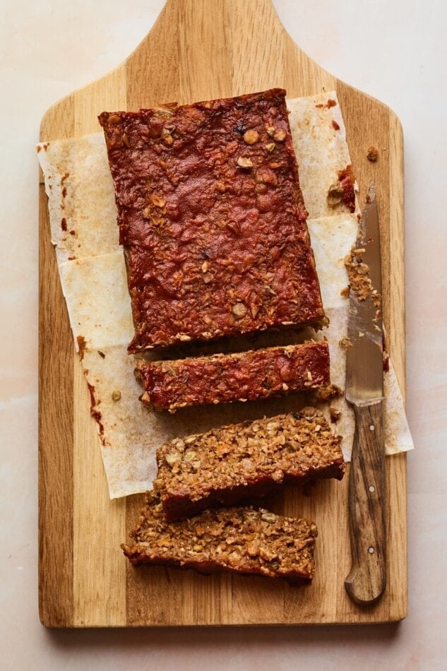 An overhead shot of a cutting board with a partially sliced lentil loaf on parchment paper. A knife lays on the cutting board perpendicular to the loaf.