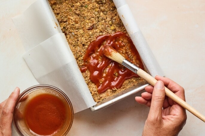 Lentil loaf mixture in a bread pan being coated with maple sweetened glaze by a hand using a small brush.