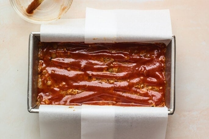 Lentil loaf coated with maple sweetened glaze in a bread pan lined with parchment paper, ready to be baked.