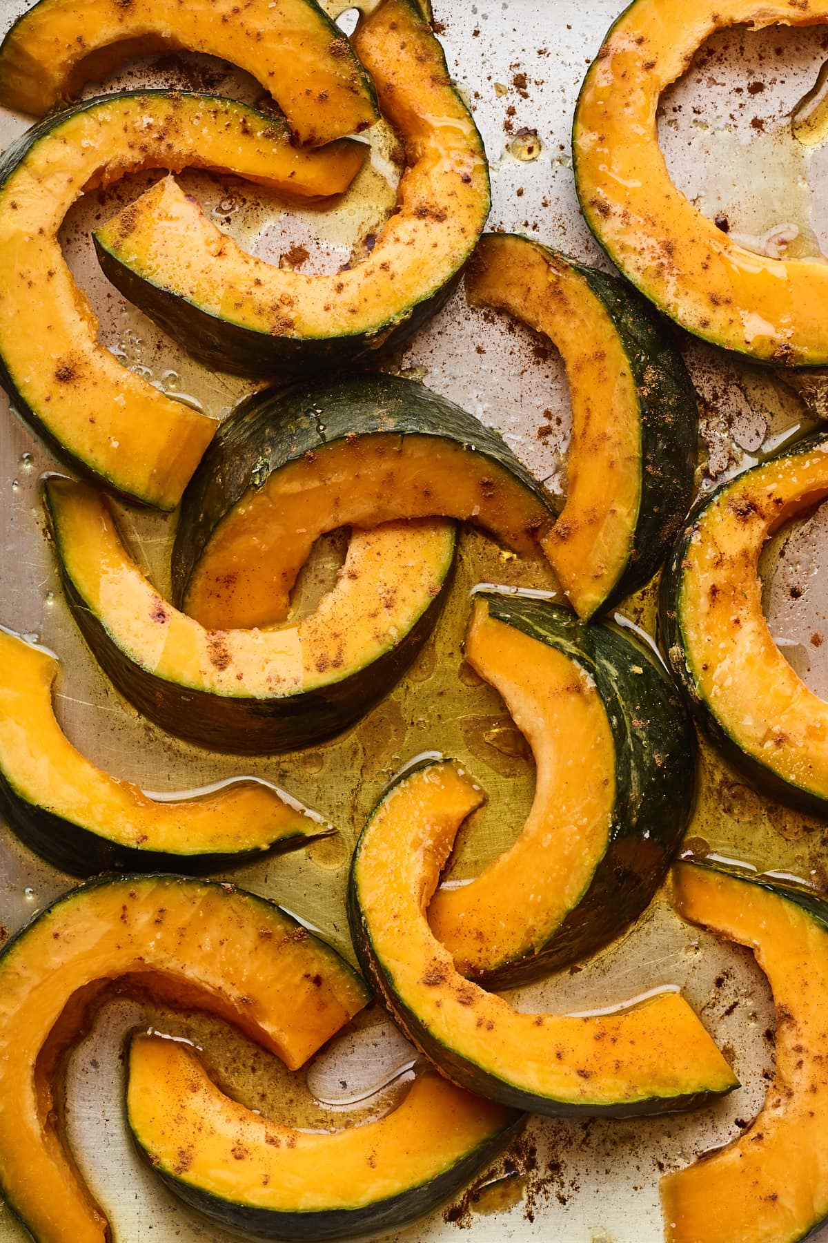 Slices of Kabocha squash covered in oil and seasoning on a sheet pan, ready to be roasted.