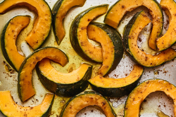 Slices of Kabocha squash covered in oil and seasoning on a sheet pan, ready to be roasted.