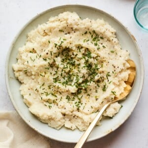A bowl of mashed cauliflower topped with chives. A serving spoon rests inside the bowl.
