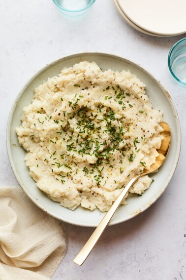 A bowl of mashed cauliflower topped with chives. A serving spoon rests inside the bowl.