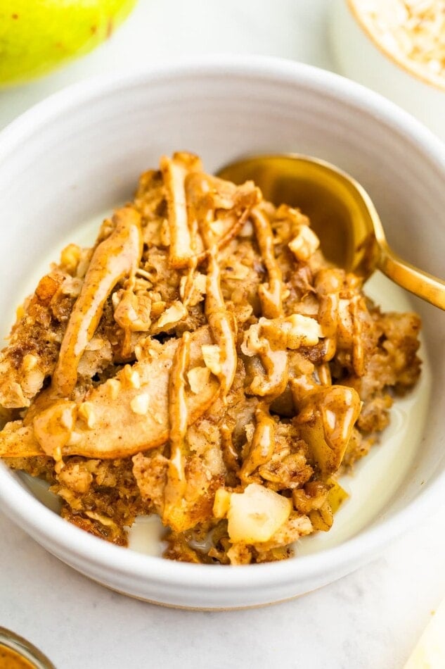 A closeup image of piece of pear baked oatmeal topped with an almond butter drizzle in a small bowl with milk and a gold spoon.