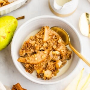 A piece of pear baked oatmeal topped with an almond butter drizzle in a small bowl with milk and a gold spoon. Ingredients are displayed around the bowl.