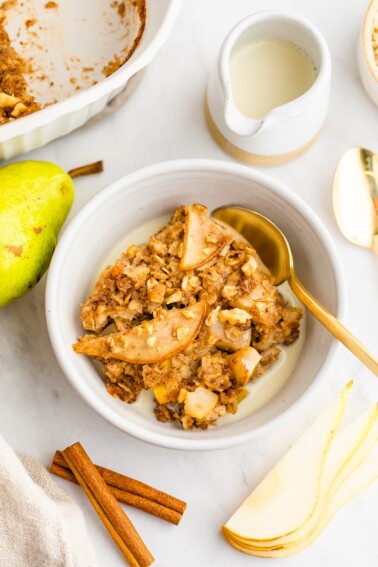A piece of pear baked oatmeal topped with an almond butter drizzle in a small bowl with milk and a gold spoon. Ingredients are displayed around the bowl.