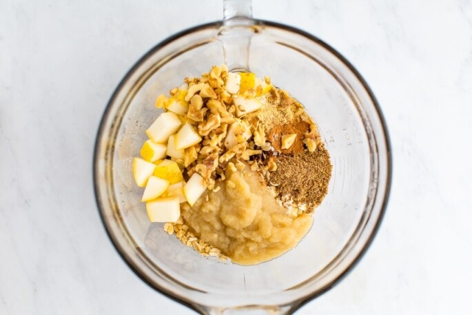 A mixing bowl with rolled oats, cinnamon, ginger, nutmeg, baking powder, salt, almond milk, applesauce, maple syrup, flaxseed, coconut oil, vanilla, walnuts and diced pears.