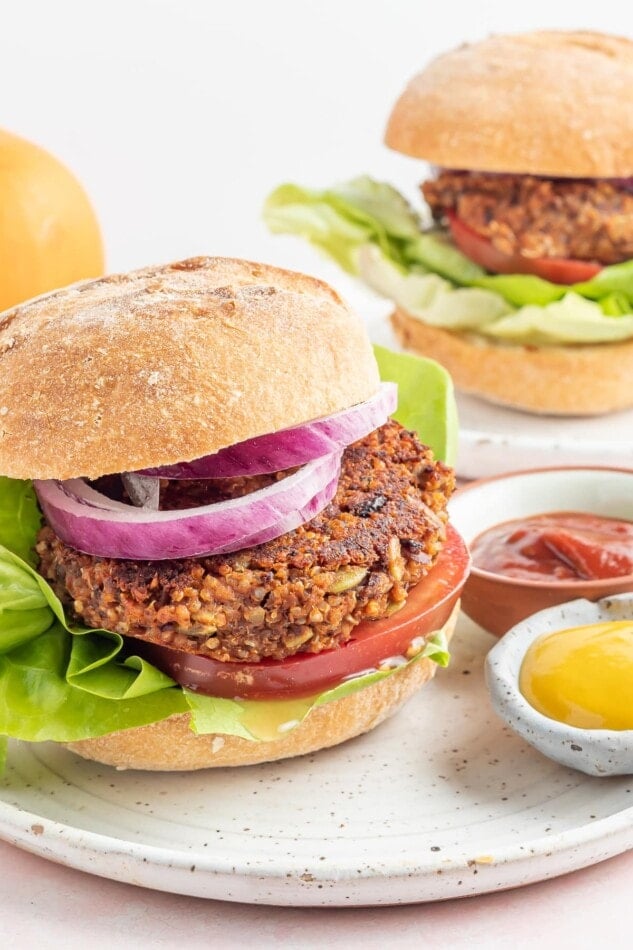 A black bean pumpkin burger on a bun with lettuce, tomato and onion. The burger is on a plate with two small ramekins of mustard and ketchup. There is another assembled black bean burger on a plate in the background.