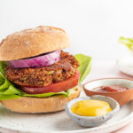 A vertical photo of a black bean pumpkin burger on a bun with lettuce, tomato and onion. The burger is on a plate with two small ramekins of mustard and ketchup.