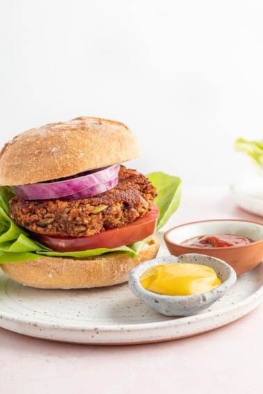 A vertical photo of a black bean pumpkin burger on a bun with lettuce, tomato and onion. The burger is on a plate with two small ramekins of mustard and ketchup.