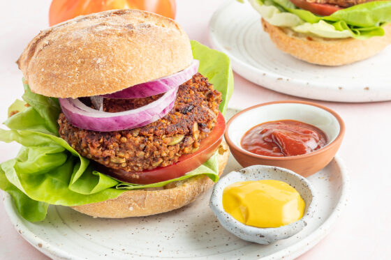 A horizontal photo of a black bean pumpkin burger on a bun with lettuce, tomato and onion. The burger is on a plate with two small ramekins of mustard and ketchup.