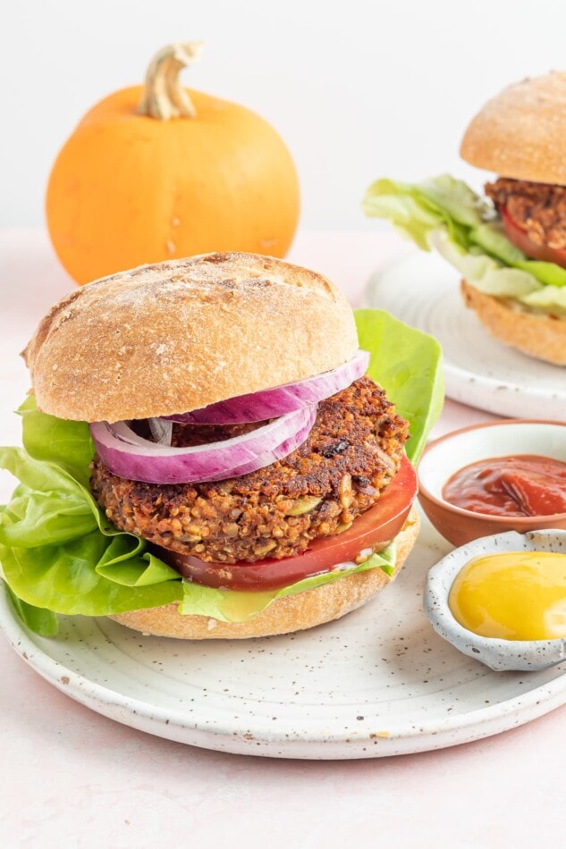 A black bean pumpkin burger on a bun with lettuce, tomato and onion. The burger is on a plate with two small ramekins of mustard and ketchup. There is a small pumpkin in the background.