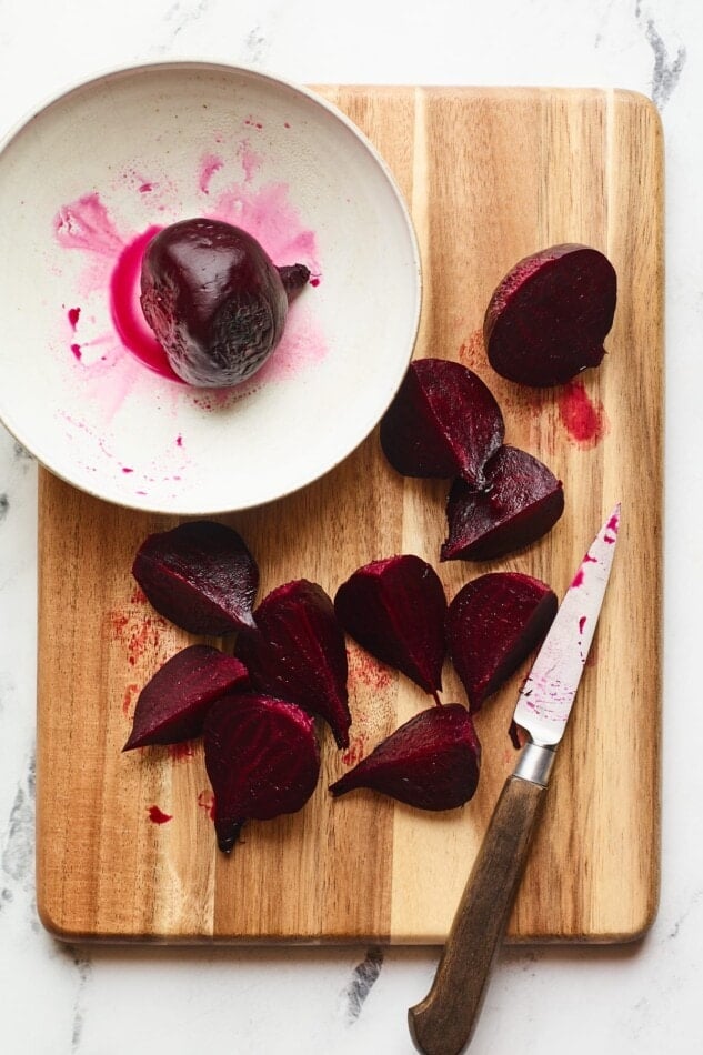 An overhead photo of a roasted beets being chopped on a wooden cutting board. A whole beet is in a shallow bowl on the cutting board.