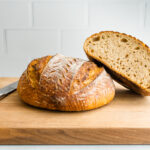 A loaf of sourdough bread on a wooden cutting board with a second loaf that has been cut in a half angled and resting on top of the other loaf. There is a bread knife to the left of the loaves on the cutting board.