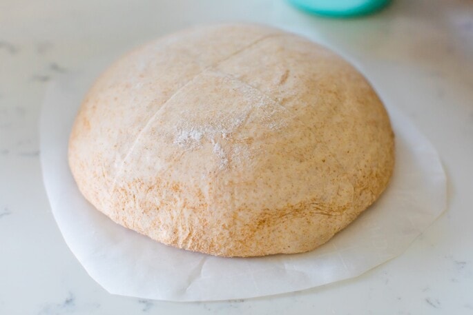 Sourdough bread dough after being removed from a banneton basket, it is resting on a piece of parchment paper on top of a counter.