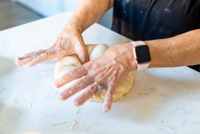 A woman is using her hands to press her thumbs into a ball of bread dough.