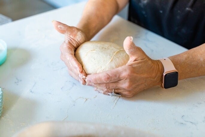 A woman is using her hands to shape a ball of bread dough.
