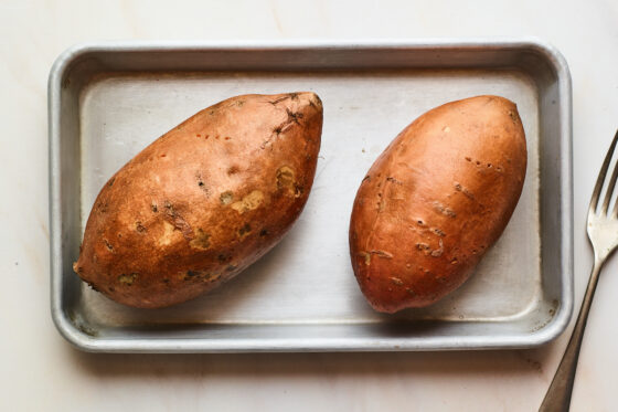Two sweet potatoes on a small baking dish with small holes poked throughout. A fork is laying next to the pan.