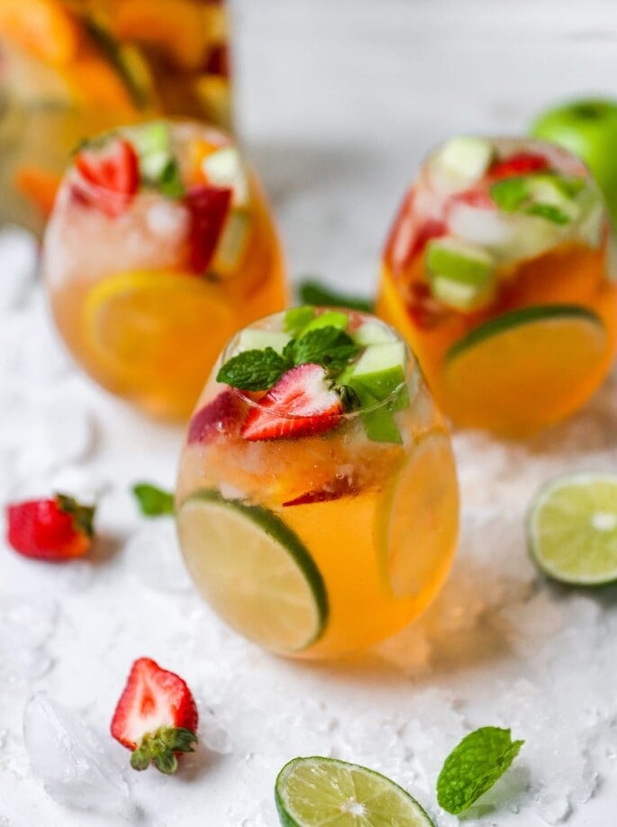 A glass of white wine sangria topped with extra strawberries and mint. Additional glasses are out of focus in the background.