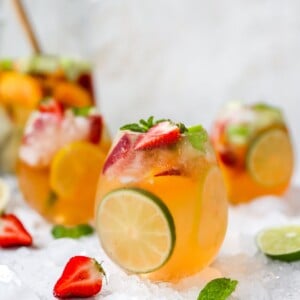 A glass of white wine sangria centered in the frame with two glasses behind it on either side out of focus. A pitcher is also out of focus in the background. There are strawberries, limes, and mint leaves scattered around the glass along with crushed ice.