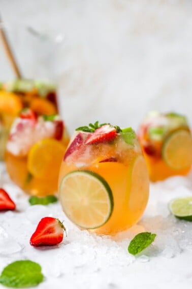 A glass of white wine sangria centered in the frame with two glasses behind it on either side out of focus. A pitcher is also out of focus in the background. There are strawberries, limes, and mint leaves scattered around the glass along with crushed ice.