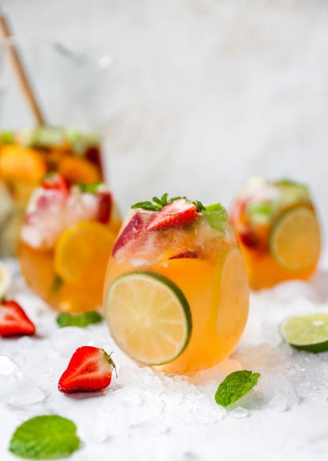 A glass of white wine sangria centered in the frame with two glasses behind it on either side out of focus. A pitcher is also out of focus in the background. There are strawberries, limes, and mint leaves scattered around the glass along with crushed ice.