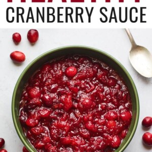 A serving dish containing cranberry sauce. A spoon rests at the top of the bowl and there are whole cranberries scattered around the bowl.