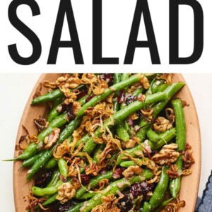 An overhead photo looking down onto a serving dish containing green bean salad topped with fried shallots. A blue linen dish cloth is laying next to the serving dish.