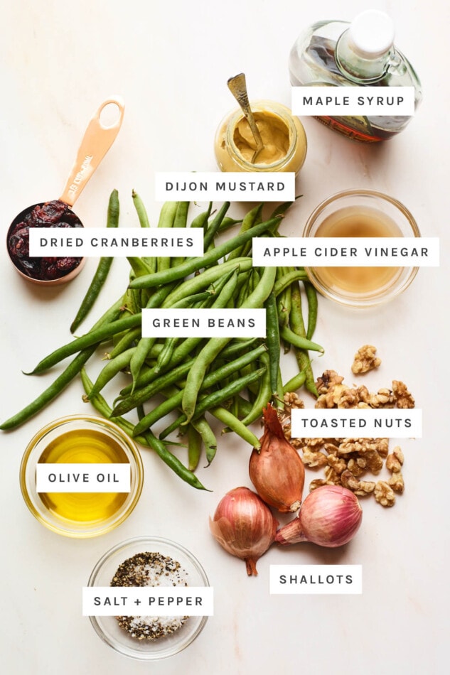 Ingredients measured out to make a green bean salad: maple syrup, dijon mustard, apple cider vinegar, dried cranberries, green beans, toasted nuts, olive oil, shallots, salt and pepper.