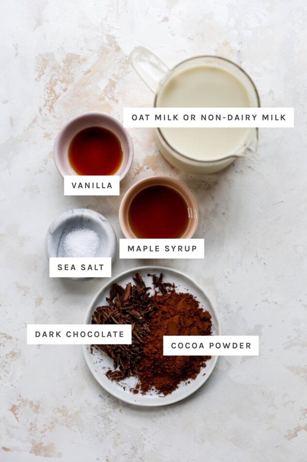 Ingredients measured out in cups and bowls to make healthy hot chocolate: oat milk, vanilla, maple syrup, sea salt, dark chocolate and cocoa powder.