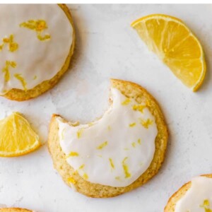 Four lemon cookies topped with glaze and lemon zest. One cookie has a bite taken out of it. Lemon slices are around the cookies.