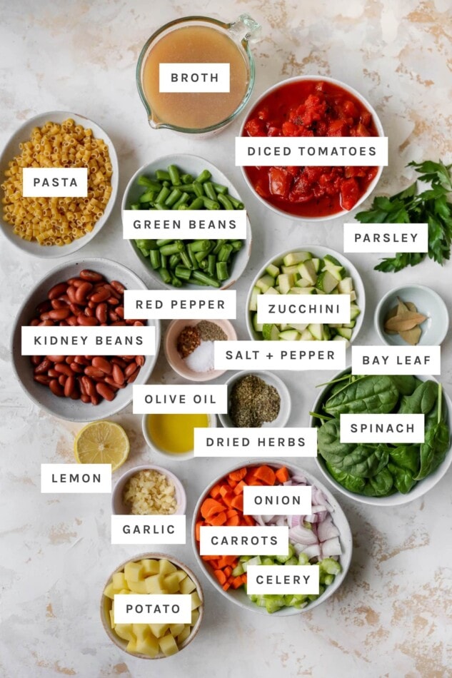 Ingredients measured out in bowls for making minestrone soup: broth, diced tomatoes, pasta, green beans, parsley, red pepper, zucchini, kidney beans, salt, pepper, bay leaf, olive oil, herbs, spinach, lemon, garlic, onion, carrots, celery and potato.