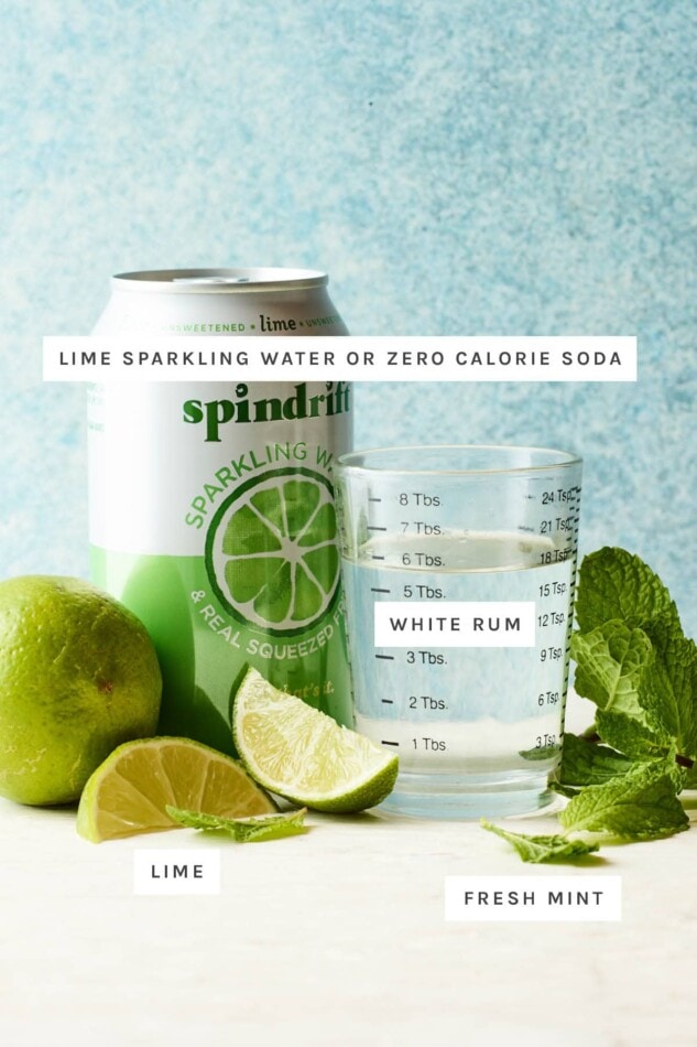 Ingredients on a table to make a Skinny Mojito: lime sparkling water, white rum, lime and fresh mint.