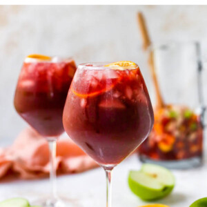 A wine glass containing red wine sangria. There is a second glass out of focus in the background along with a pitcher.