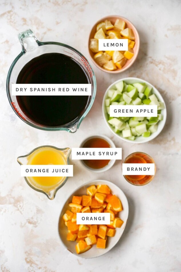 Ingredients measured out to make red sangria: chopped lemon, chopped green apple, dry spanish red win, maple syrup, brandy, orange juice and chopped orange.