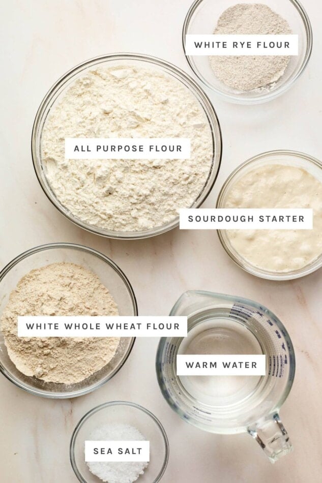 Ingredients measured out in glass bowls to make sourdough bread: white rye flour, all purpose flour, sourdough starter, white whole wheat flour, warm water and sea salt.