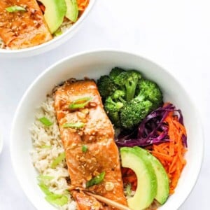 An overhead photo looking at a bowl of rice topped with a filet of teriyaki salmon, broccoli, shredded carrots, shredded cabbage, green onion and sliced avocado. There are two chopsticks resting on the bowl.