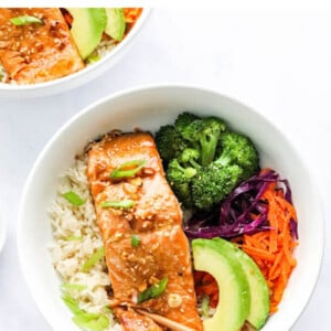 An overhead photo looking at a bowl of rice topped with a filet of teriyaki salmon, broccoli, shredded carrots, shredded cabbage, green onion and sliced avocado. There are two chopsticks resting on the bowl.