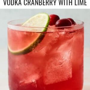 A closeup photo of a short glass filled with vodka cranberry over ice and topped with a slice of lime and a few whole cranberries.
