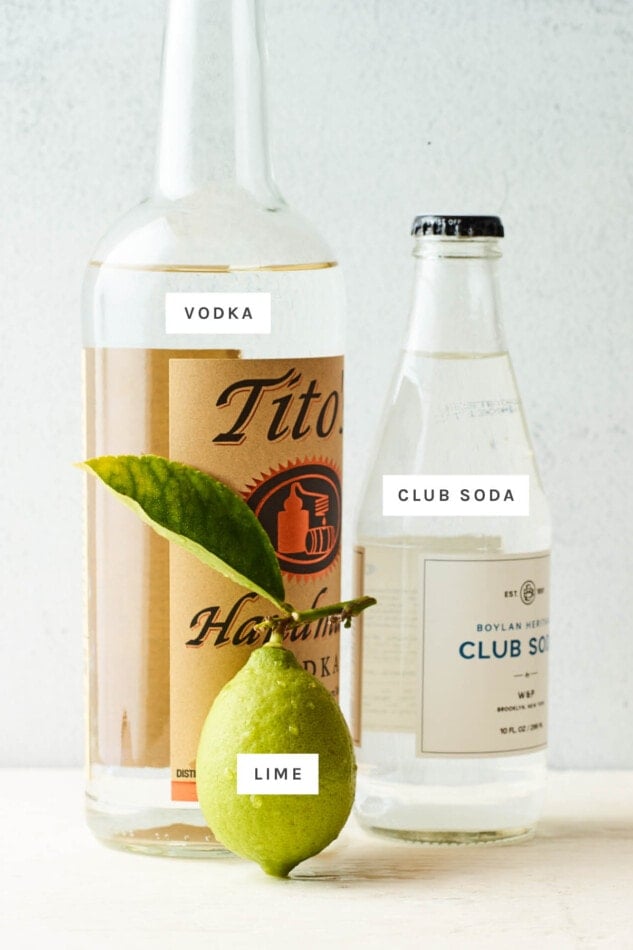 Ingredients on a table to make a vodka soda: bottle of Tito's vodka, bottle of club soda and a whole lime.