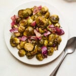 A large serving of roasted brussels sprouts and red onion on a white plate with a metal spoon to the side.