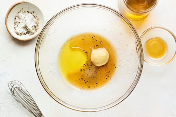 A glass bowl with apple cider vinegar, oil, maple syrup, mustard, salt and pepper ready to be whisked together. A whisk is laying next to the bowl.